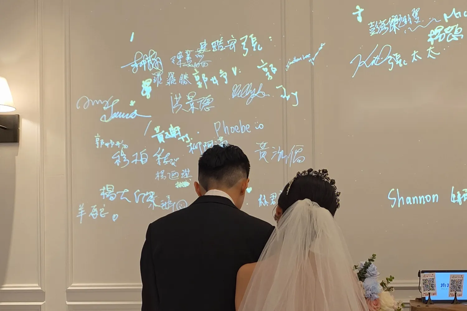 A couple, seen from behind, stand before a wall covered in their guests’ projected signatures
