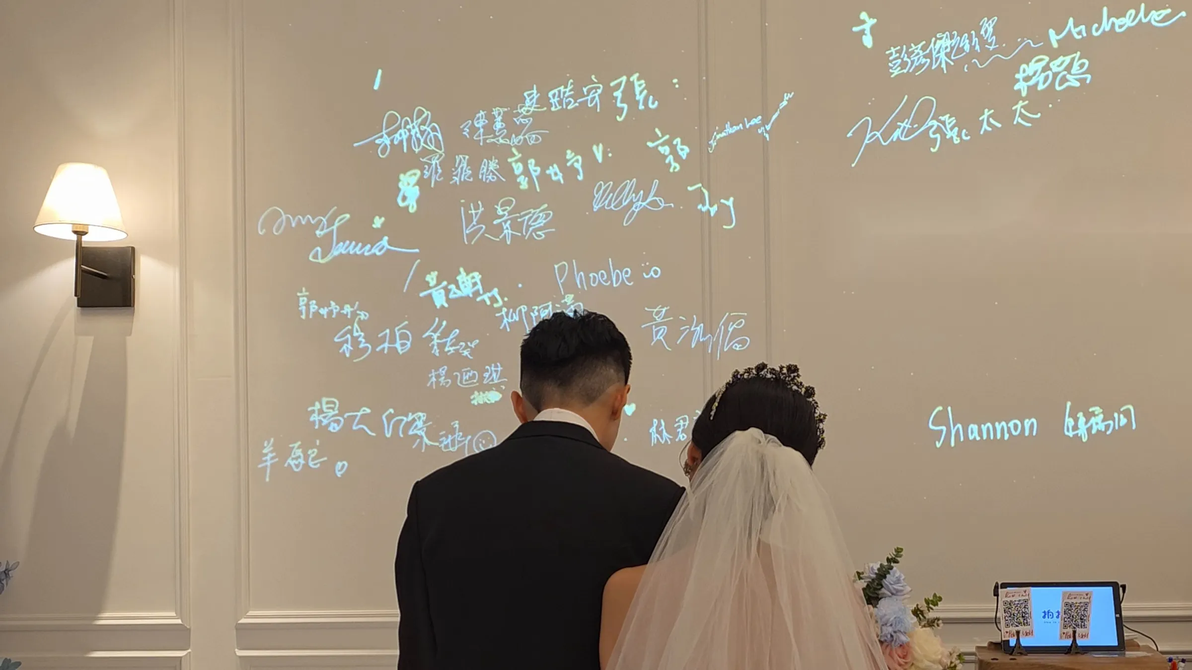 A couple, seen from behind, stand before a wall covered in their guests’ projected signatures